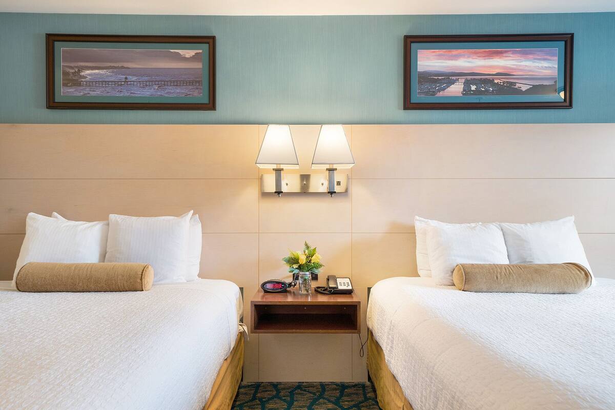 A hotel room with two beds, a nightstand with a lamp, a phone, and a small vase with flowers.
