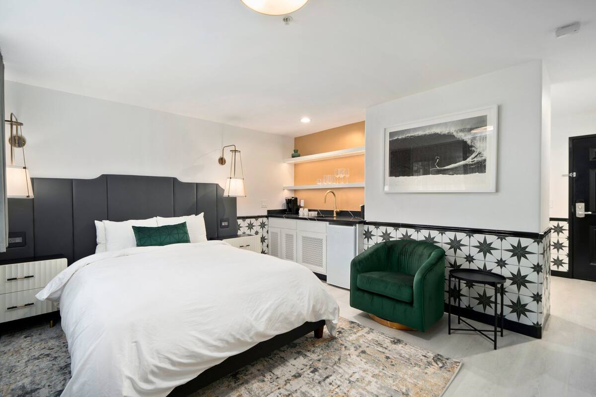 A modern hotel room with a large bed, a green armchair, a small table, a kitchen area, and a framed artwork on the wall.