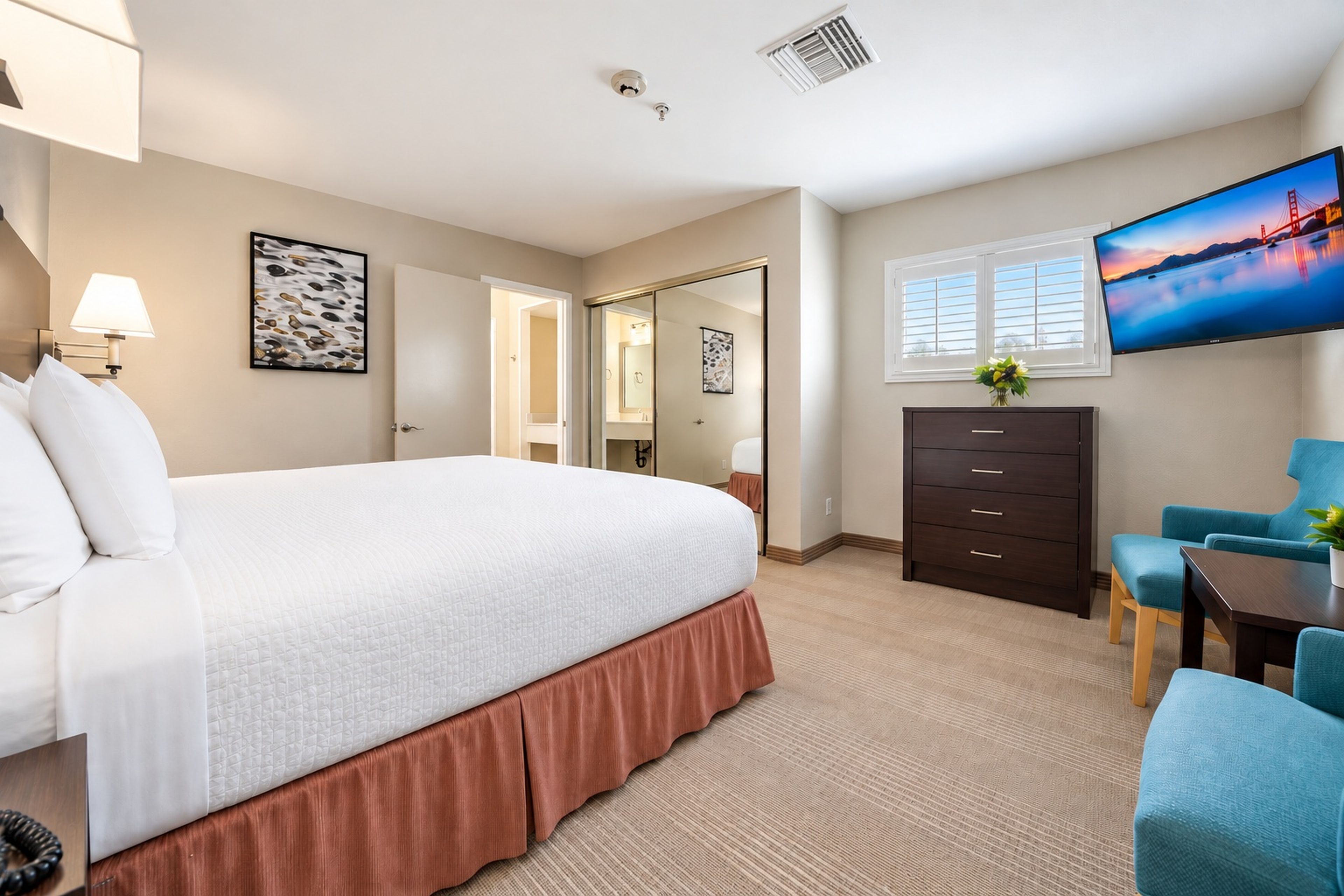 A hotel room with a bed, a dresser, a desk with a chair, a TV on the wall, and a window with a view.