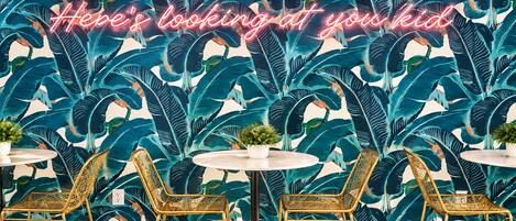 A room with a vibrant tropical leaf wallpaper, three round tables with chairs, and a neon sign.
