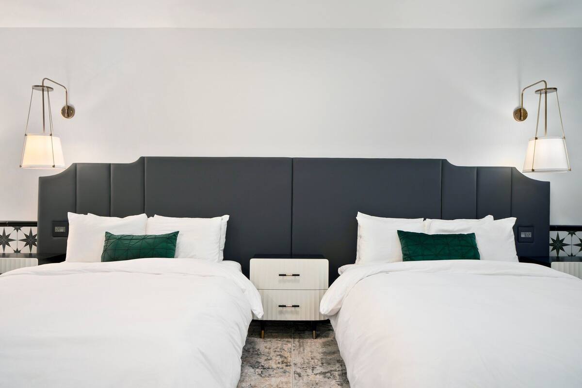 A hotel room with a dark headboard, two beds with white bedding, a small bedside table, and two wall-mounted lamps.