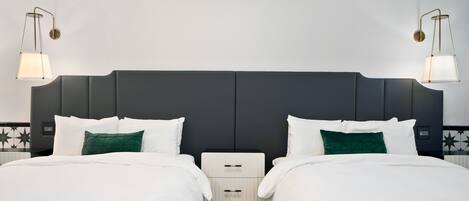 A hotel room with a dark headboard, two beds with white bedding, a small bedside table, and two wall-mounted lamps.