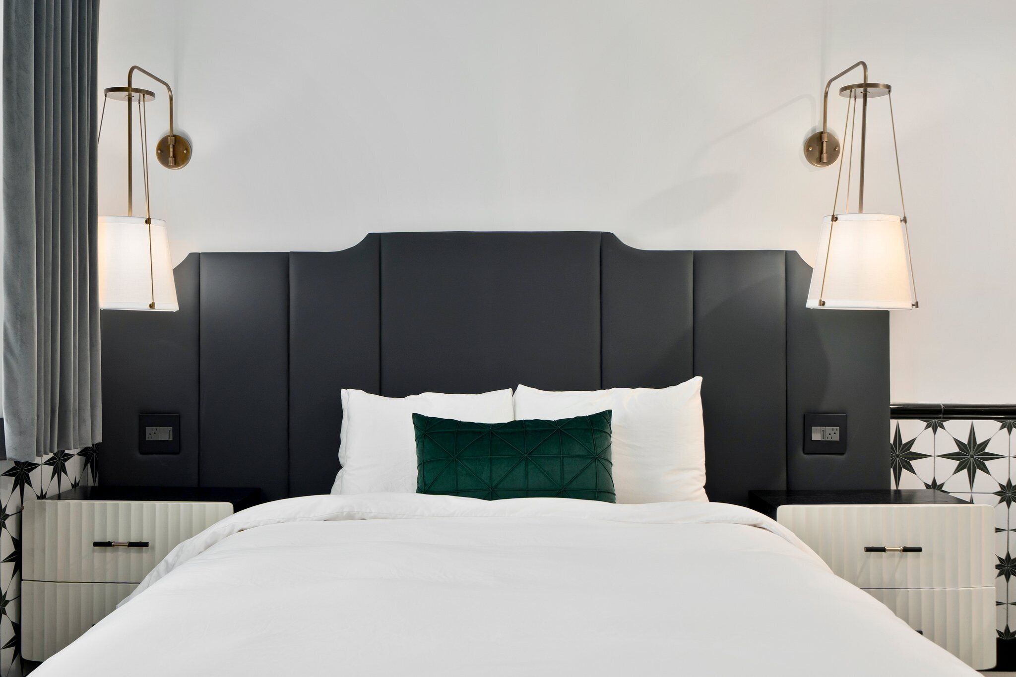 Una habitación de hotel moderna con una cama grande, cabecero oscuro, mesitas de noche y lámparas fijadas a la pared.