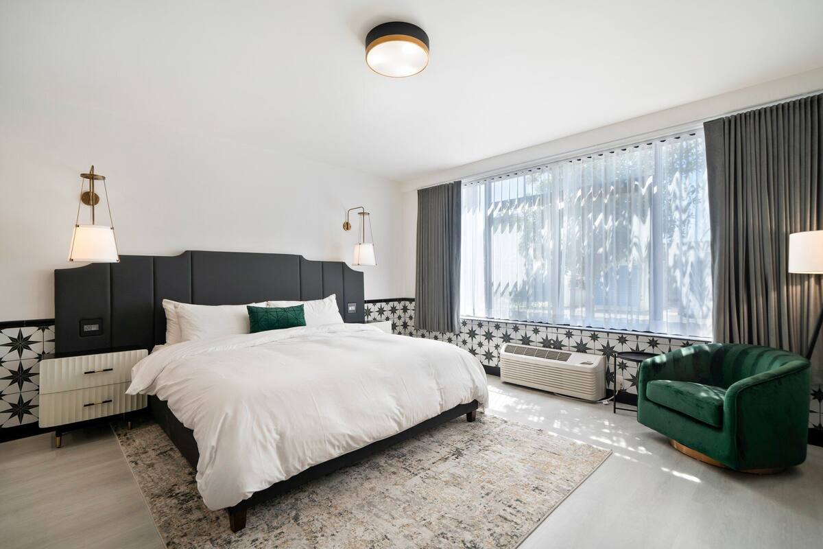 A modern bedroom with a large bed, a green armchair, and a window with curtains.