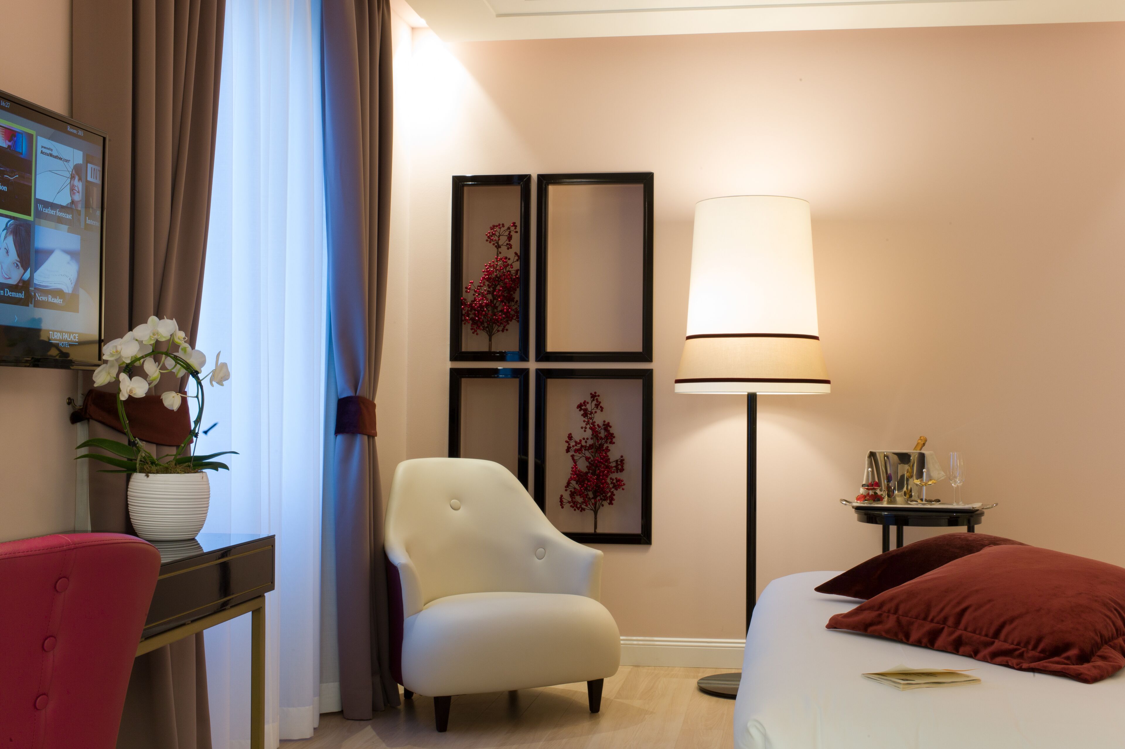 Ein Hotelzimmer mit einem Bett, einem Schreibtisch mit Blumen in einer Vase, einem Sessel, einer Lampe und gerahmten Bildern an der Wand.