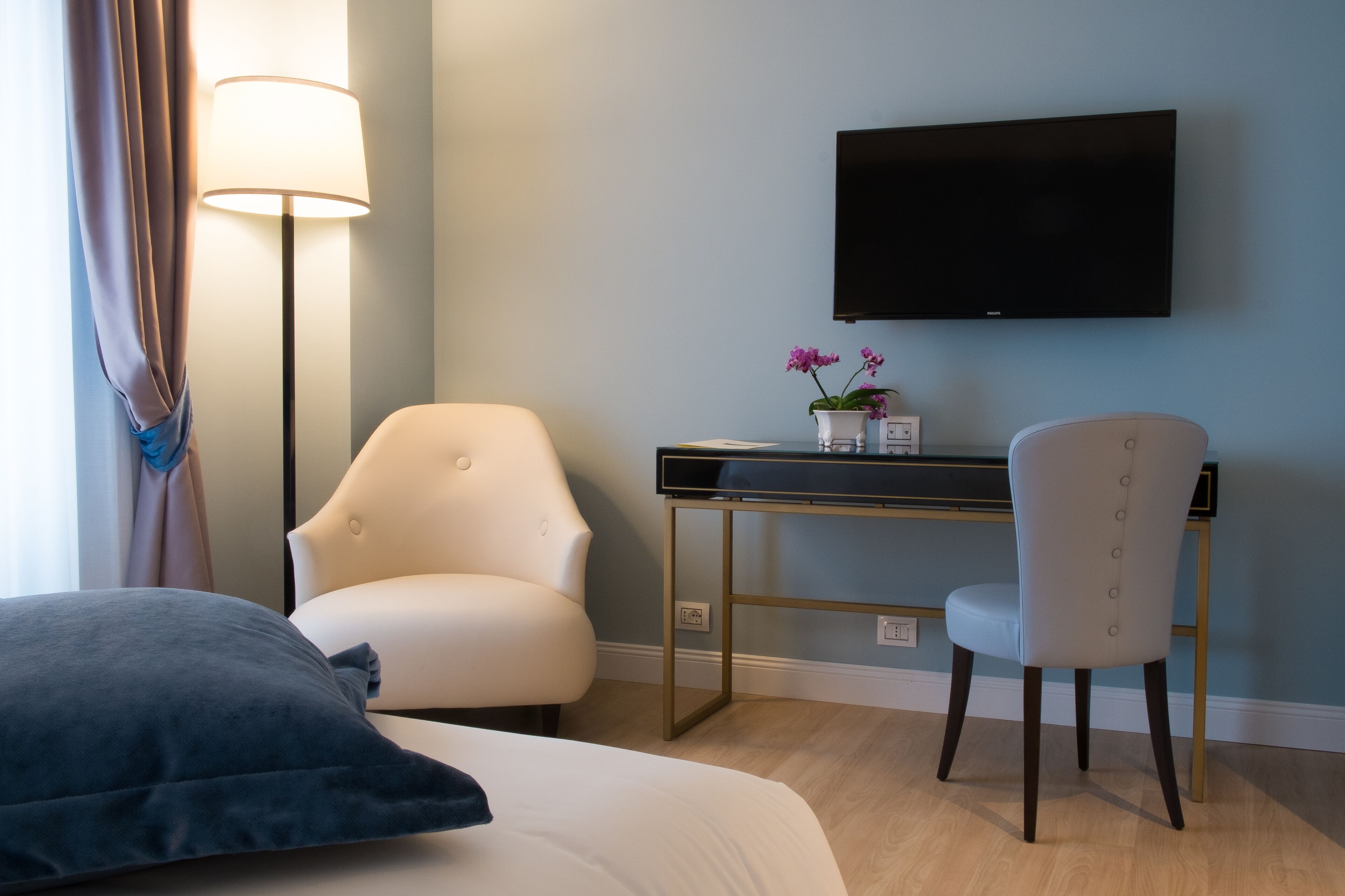 Habitación de hotel con cama, escritorio con televisor de pantalla plana, dos sillas, lámpara y pequeña mesa con flores.