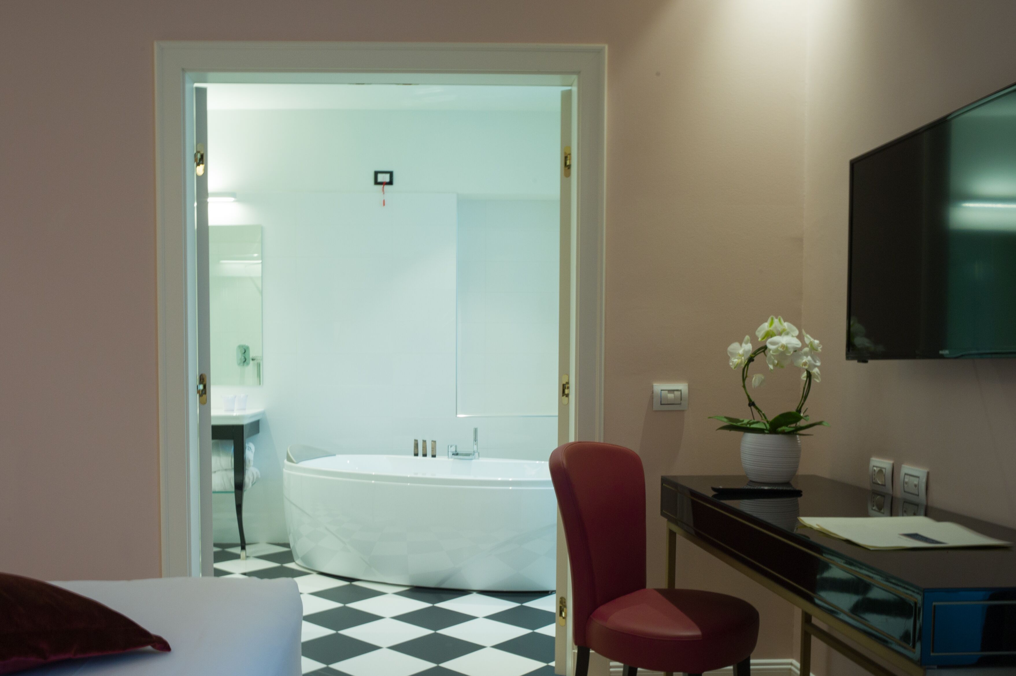 Une chambre d’hôtel avec un sol à carreaux, une chaise rouge, un bureau avec un téléviseur à écran plat, une plante en pot et une baignoire.