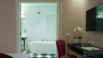 A hotel room with a checkered floor, a red chair, a desk with a flat-screen TV, a potted plant, and a bathtub.