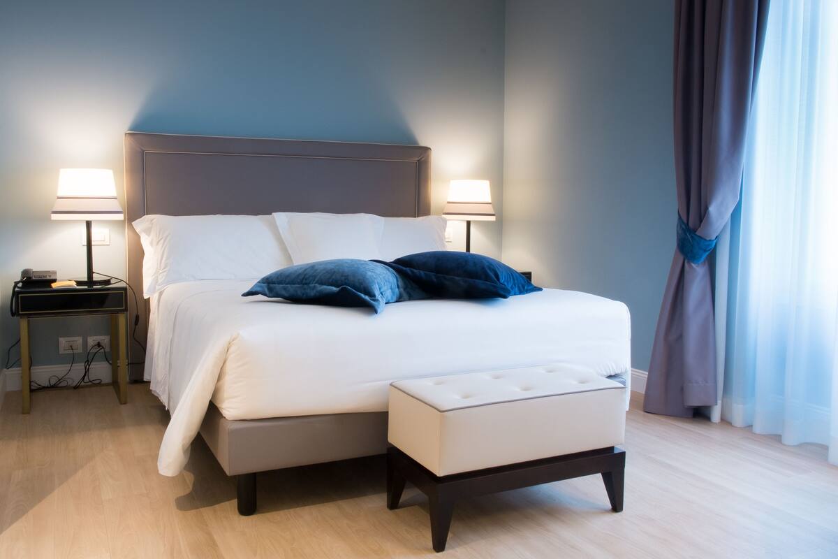 Un lit bien fait, avec des oreillers blancs et bleus, une table de chevet avec une lampe, et un petit banc au pied du lit.