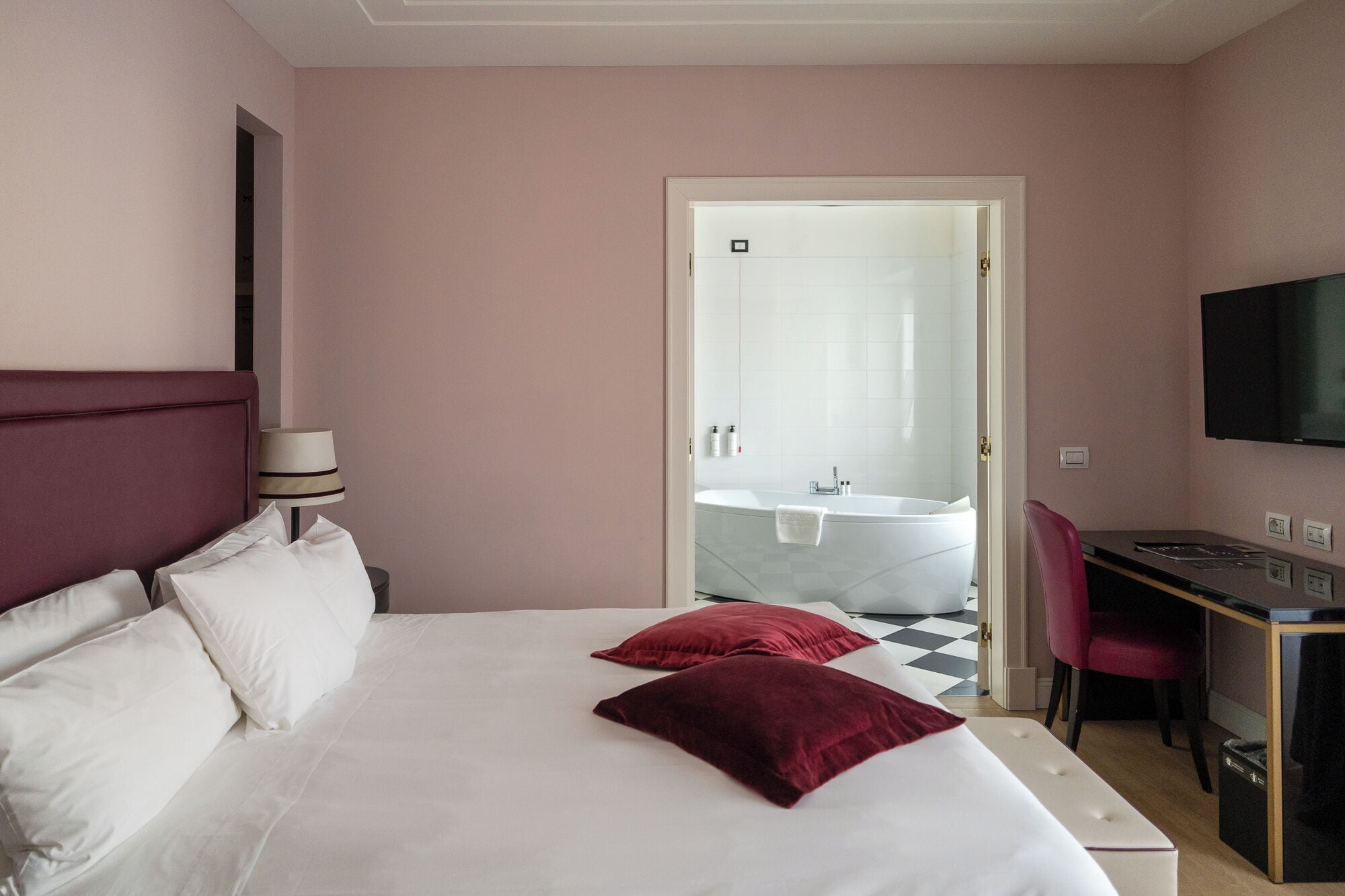 A hotel room with a bed, a desk, a chair, a TV, and a bathroom visible through an open door.