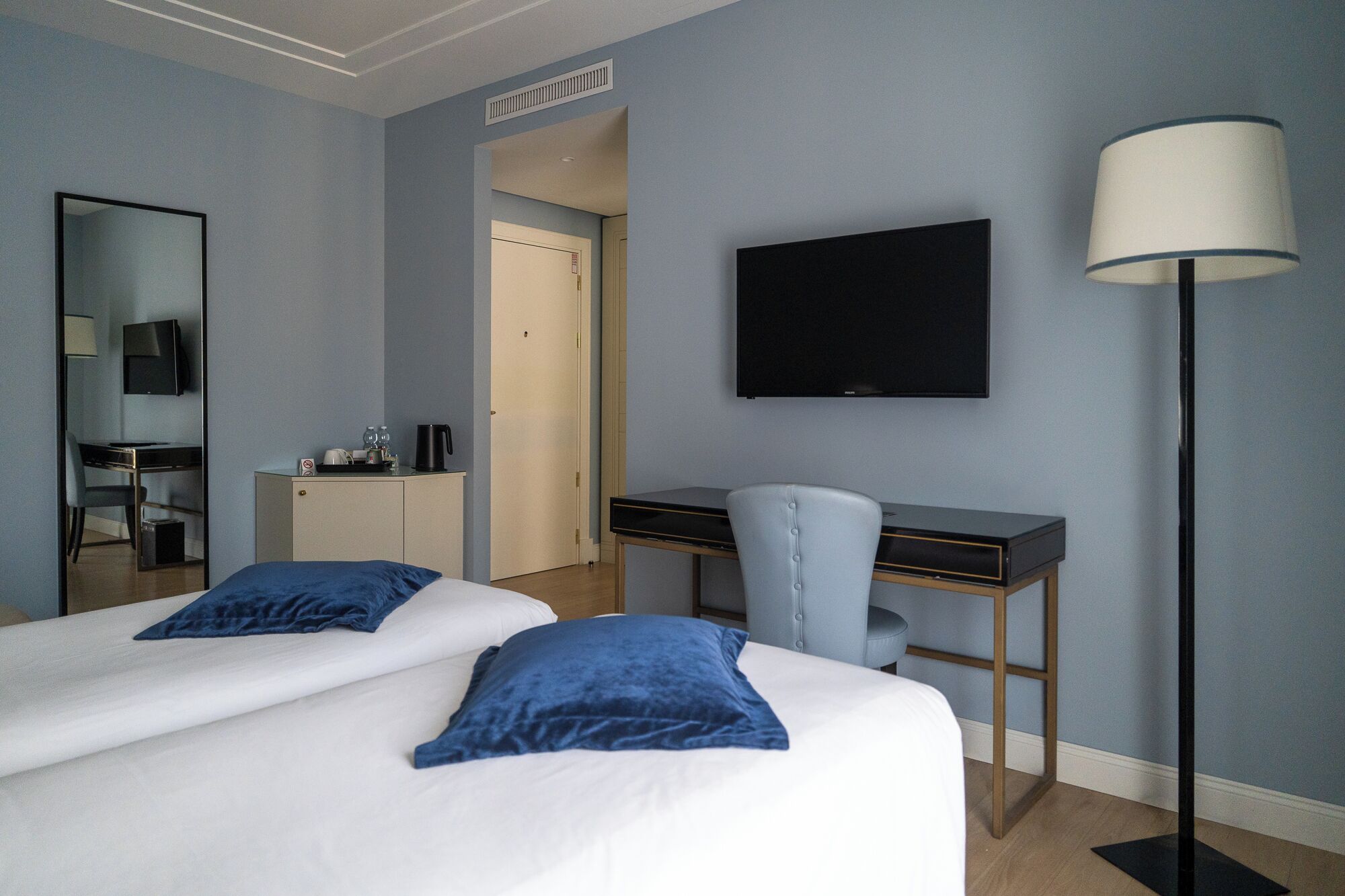 A hotel room with two beds, a desk, a chair, a TV, and a lamp.