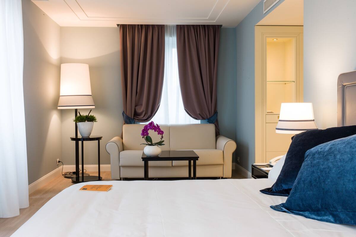 Une chambre d’hôtel avec un lit, un canapé, une petite table avec une plante, une lampe et une fenêtre avec des rideaux.