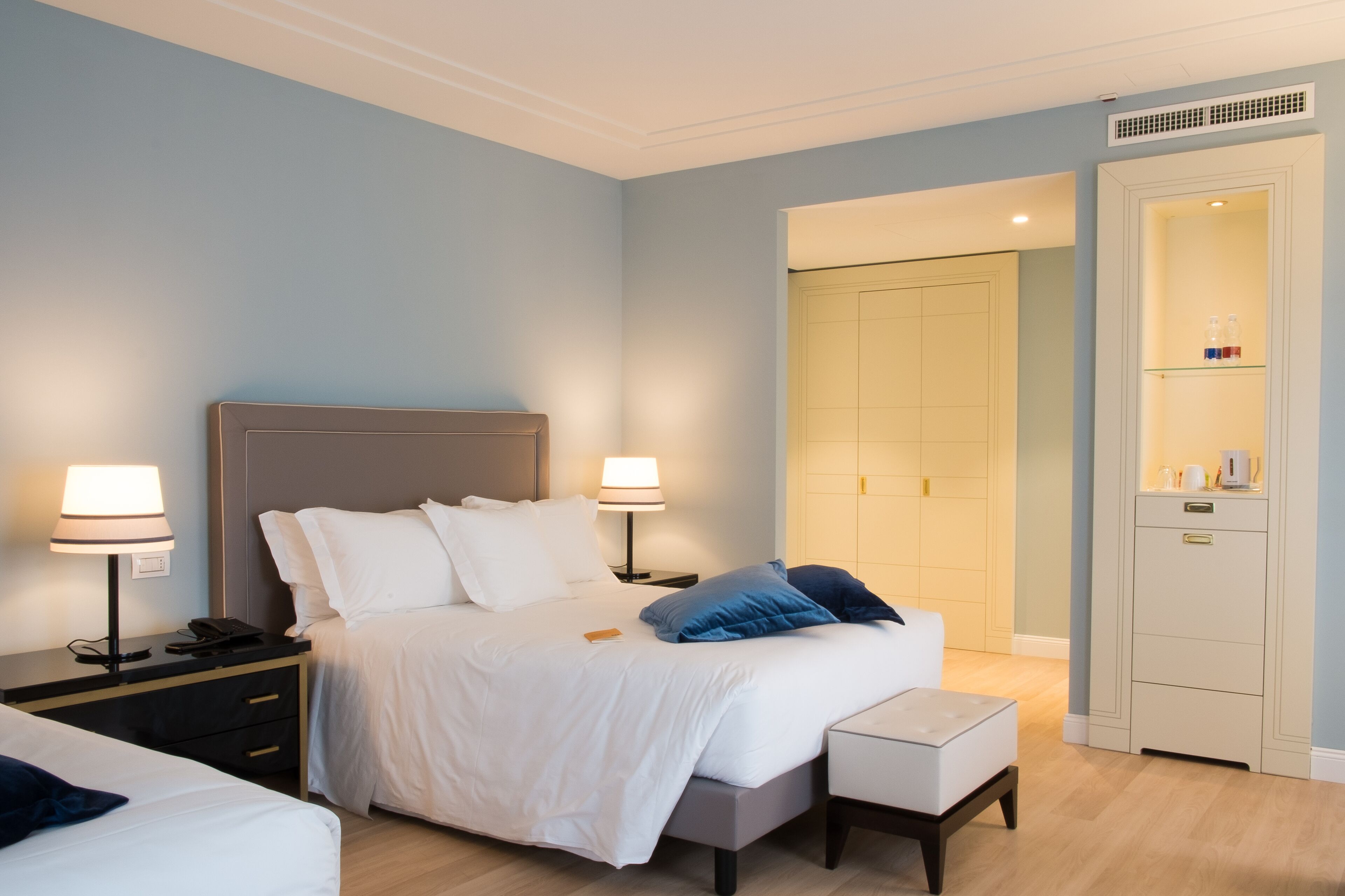 Une chambre d’hôtel avec un grand lit, des tables de chevet avec des lampes, une petite table de chevet avec un téléphone, un banc et une armoire.