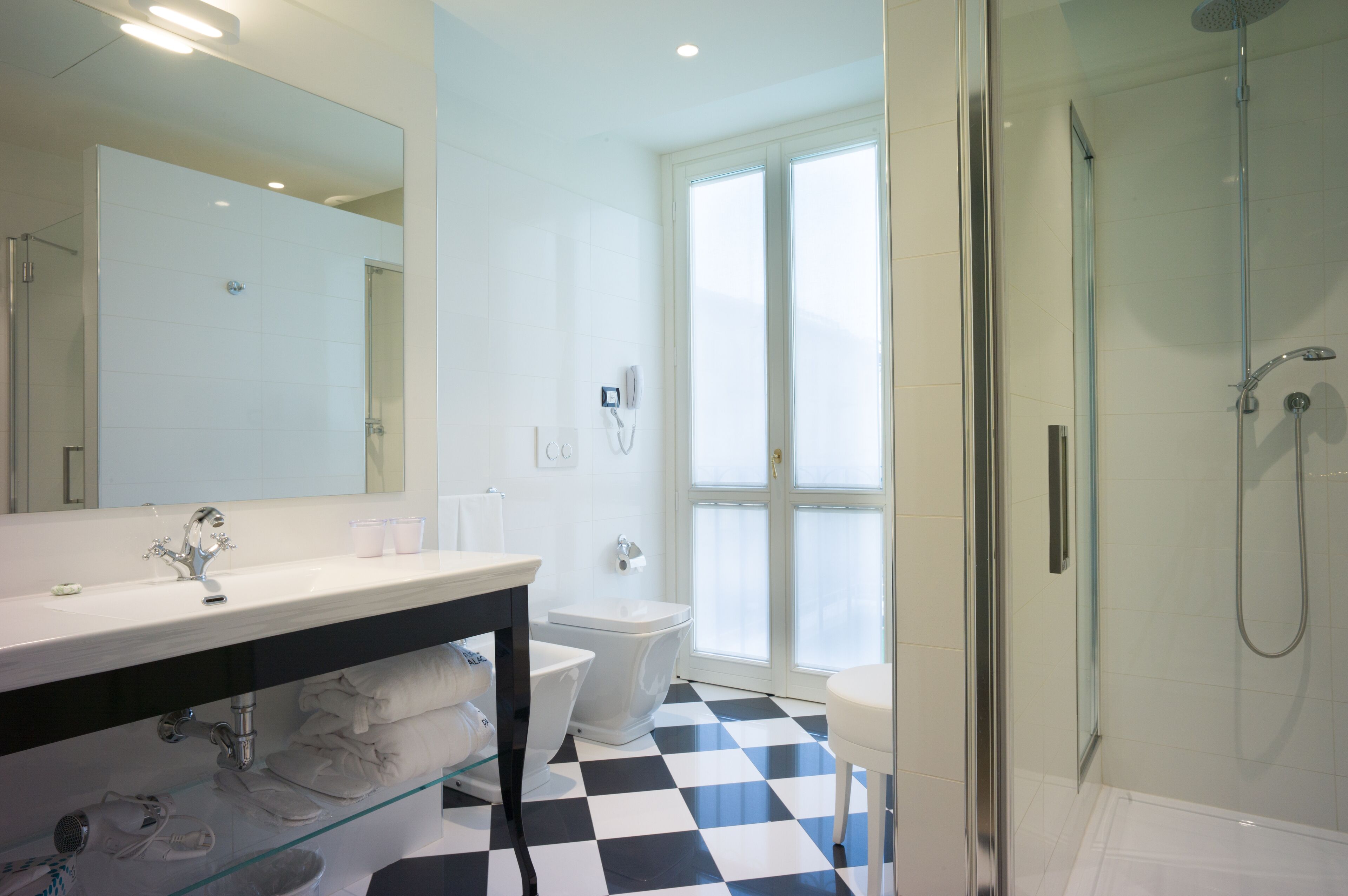 Un bagno moderno con un grande specchio, sanitari bianchi, cabina doccia in vetro e pavimento a scacchi.