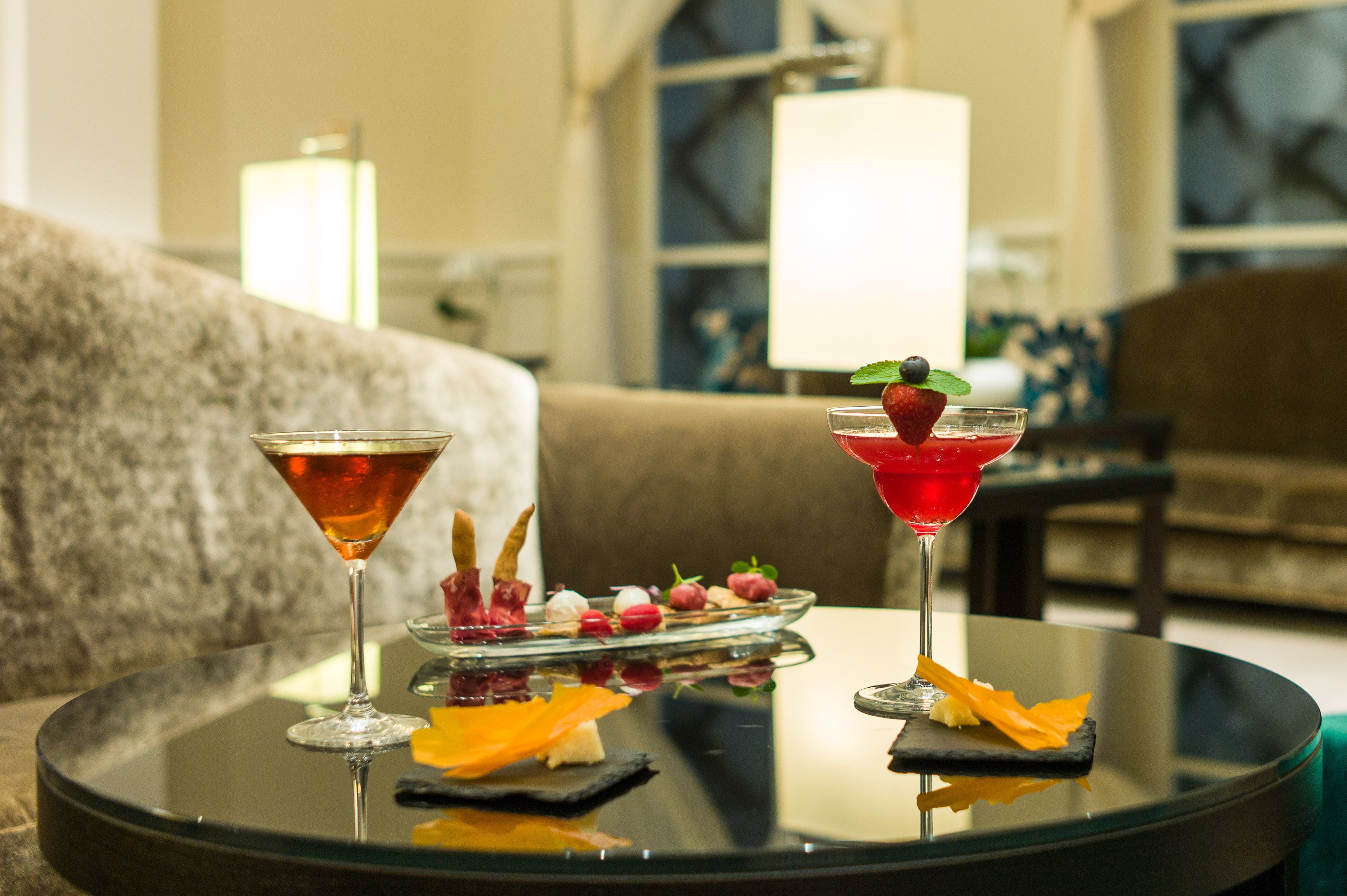 Un tavolo rotondo in vetro con due bicchieri da martini, un cocktail decorato con frutta e un piccolo piatto di stuzzichini.