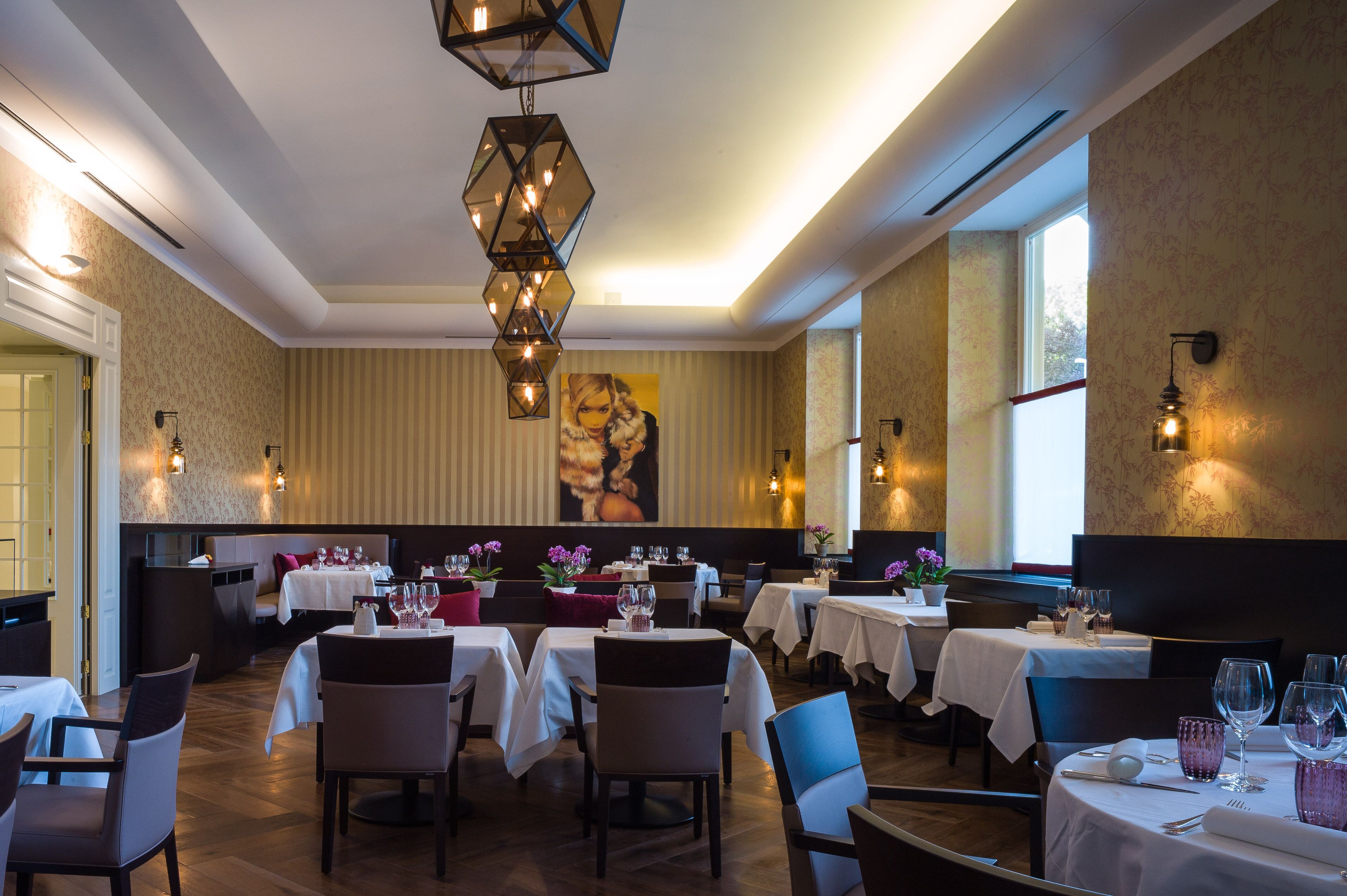 A restaurant with multiple tables set for dining, a large painting on the wall, and pendant lighting.