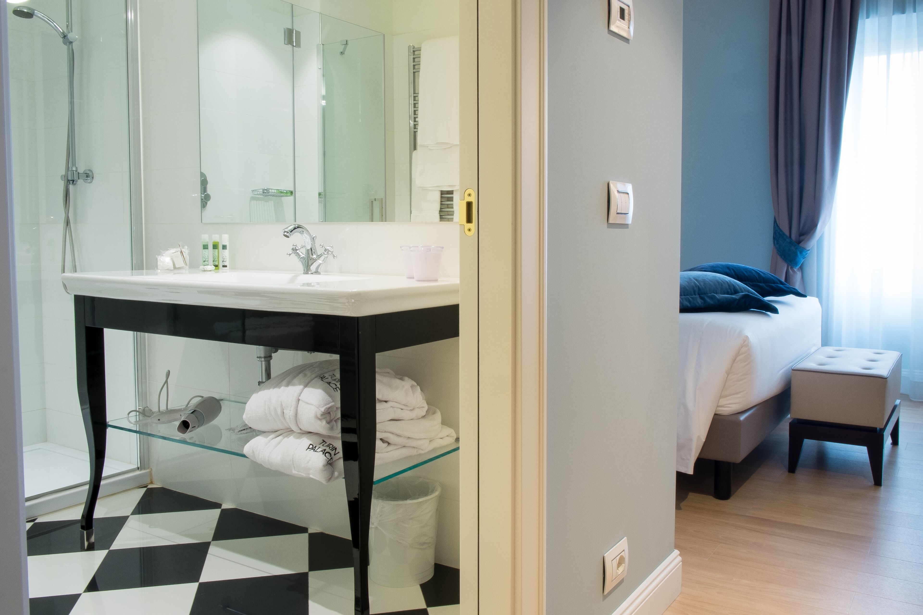 Habitación de hotel con cabina de ducha de vidrio, lavamanos blanco, cama con almohada azul y un pequeño otoman.