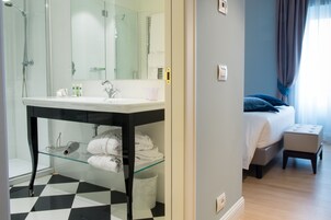 Una camera d'albergo con box doccia in vetro, un lavandino bianco, un letto con cuscino blu e un piccolo poggiapiedi.