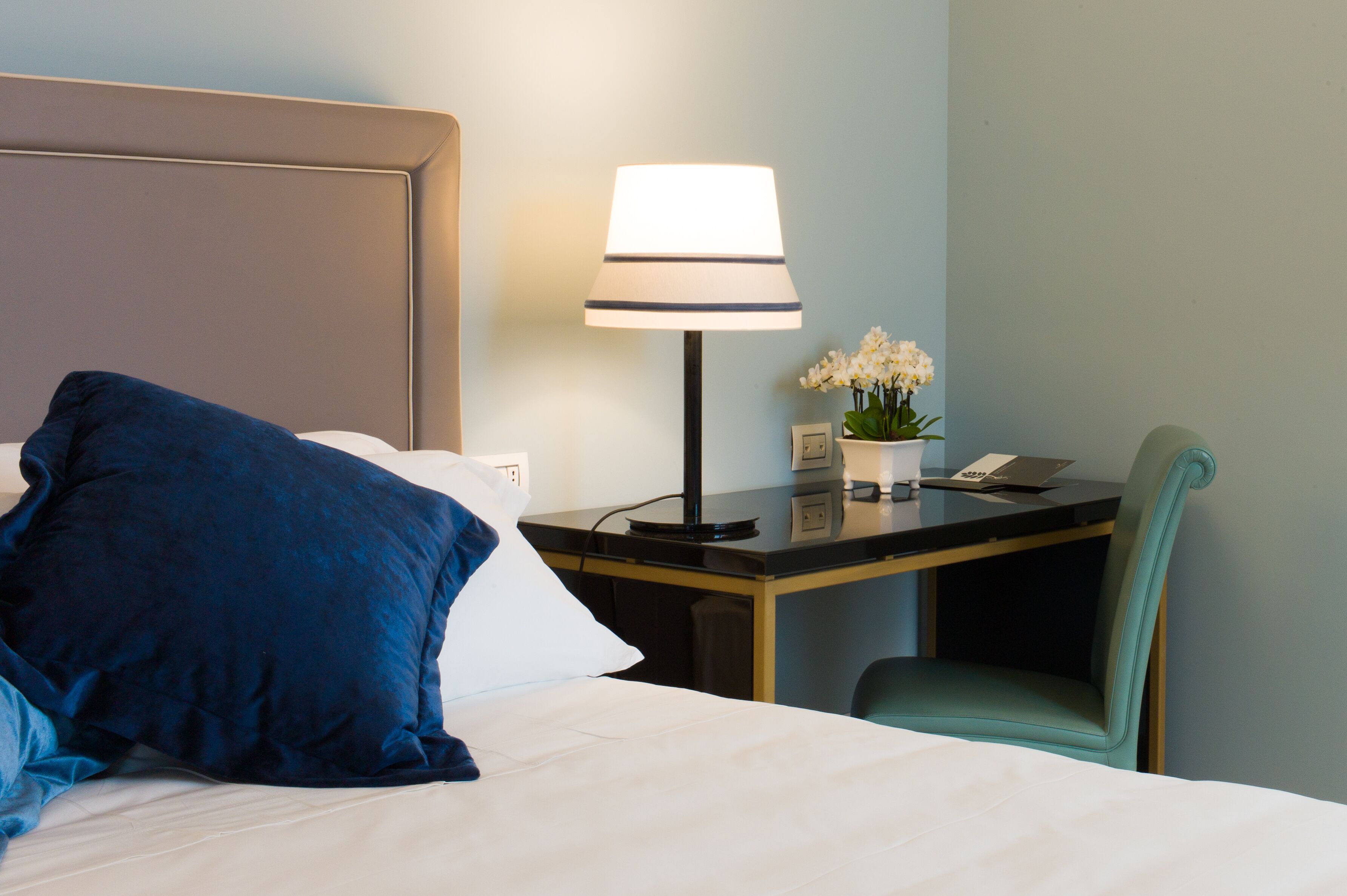 Quarto de hotel com cama, escrivaninha com luminária, cadeira e mesa pequena com planta e telefone.