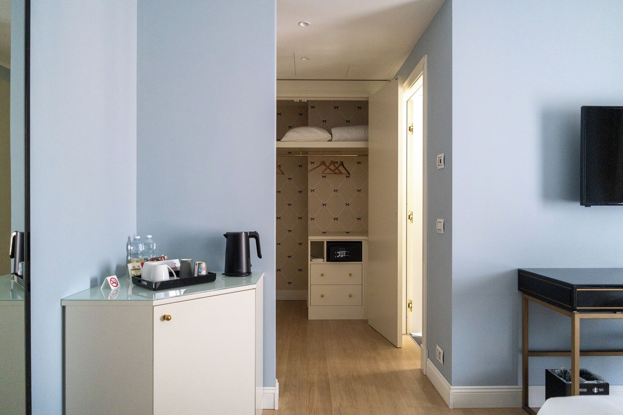 A compact hotel room with a kitchenette, a bed with a headboard, a wardrobe, and a television on the wall.