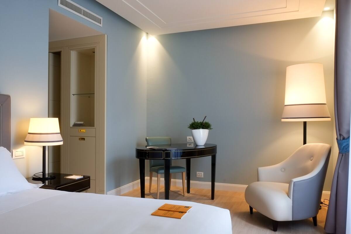 Une chambre d’hôtel avec un lit, un bureau, une chaise, une lampe et une plante en pot.