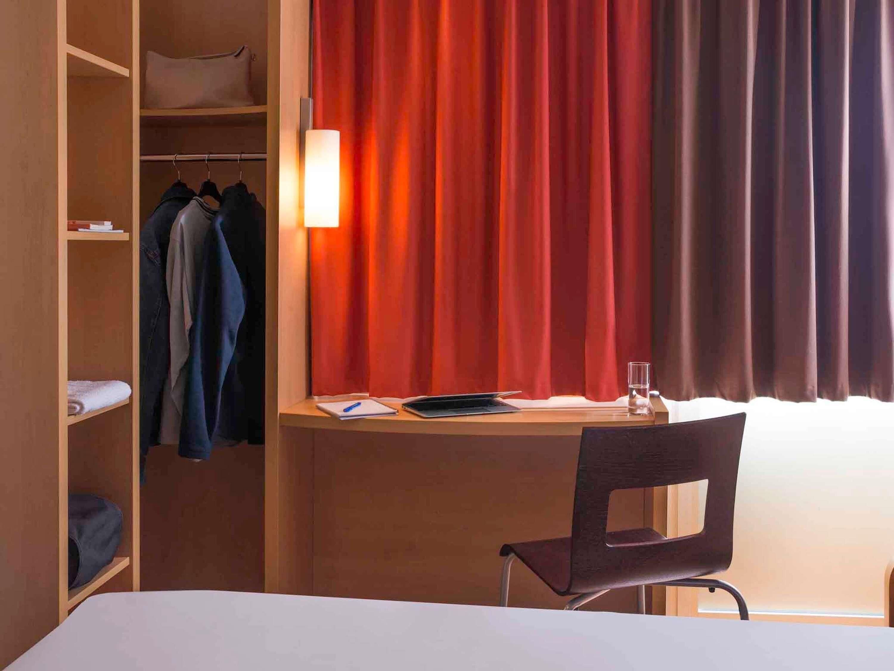 Une chambre d’hôtel comprenant un lit, un bureau avec une chaise, une armoire et une fenêtre avec des rideaux.