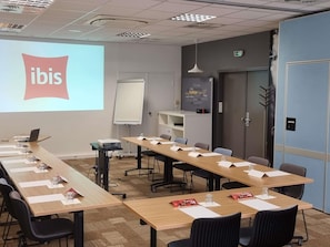 Een vergaderruimte met een projectiescherm waarop het 'ibis'-logo staat, een whiteboard, een plank met boeken en een deur met een vluchtsignaal.