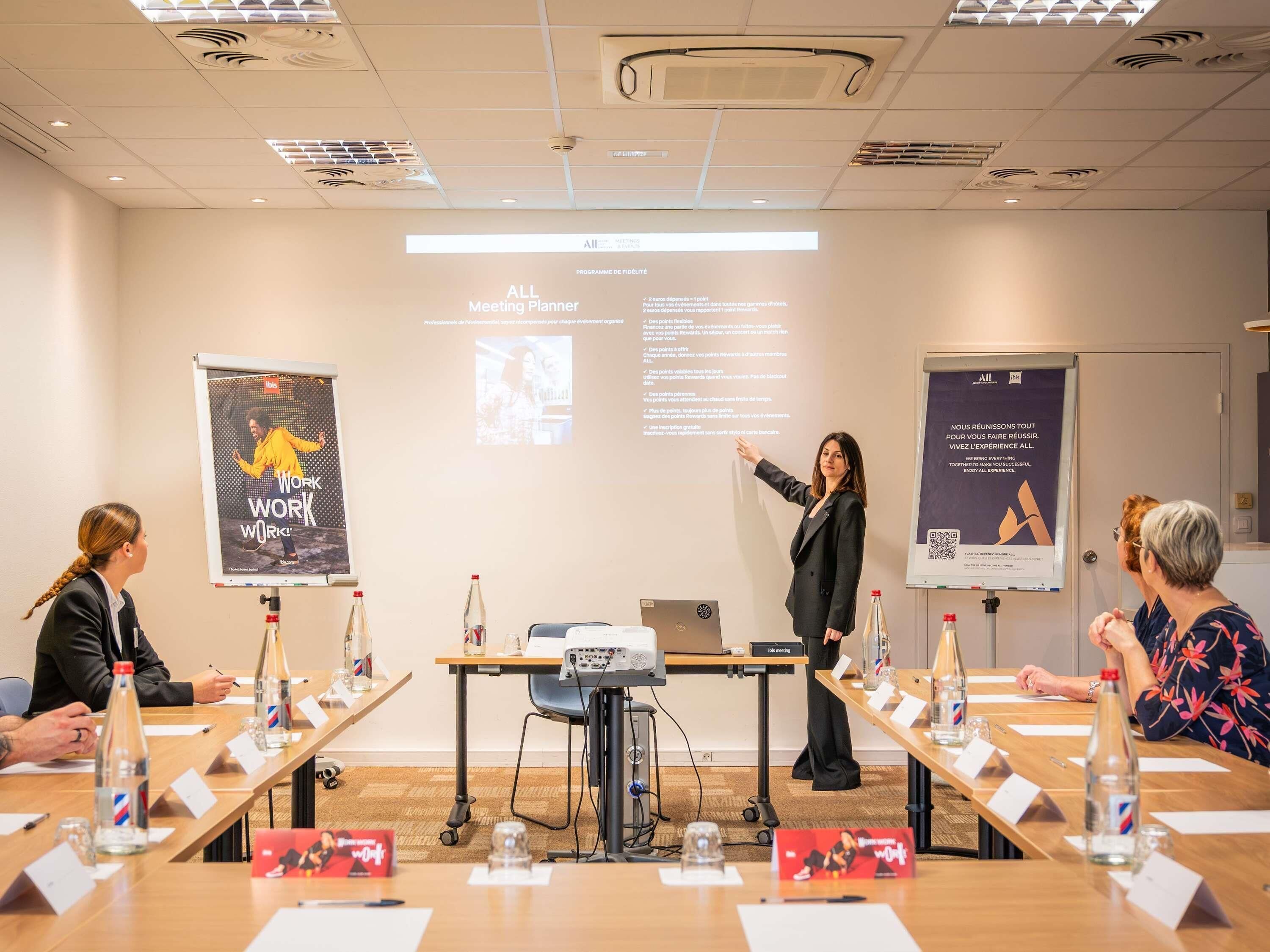 Ett konferensrum med en projektorduk, presentationsbilder och en talare som vänder sig till publiken.