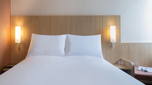 Un lit bien fait, avec du linge de lit blanc, deux lampes de chevet fixées au mur, et une petite table de chevet sur laquelle se trouvent un verre d’eau et une paire de lunettes de soleil.