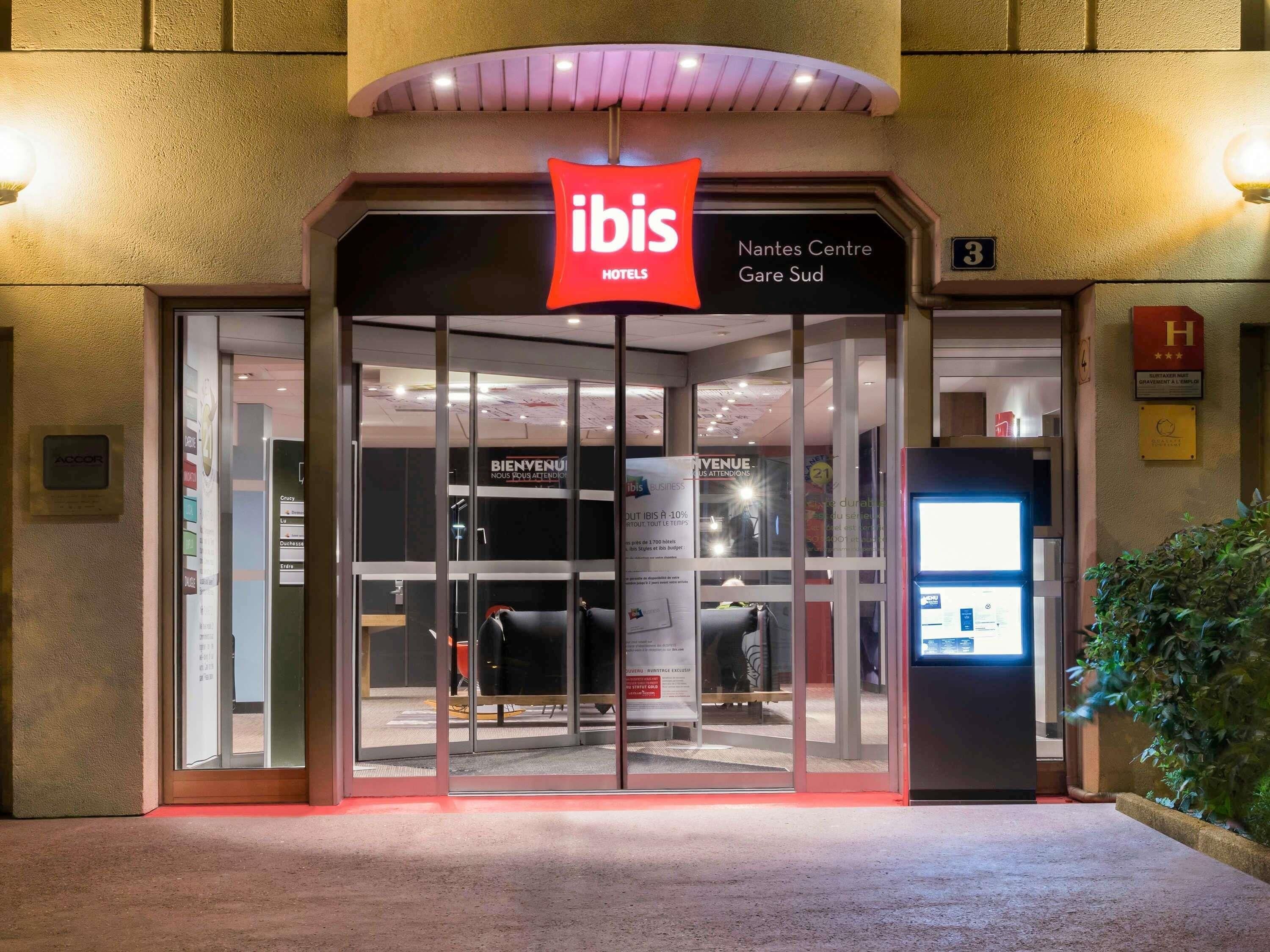La entrada a un hotel ibis con fachada de vidrio, un letrero rojo y una pantalla digital.
