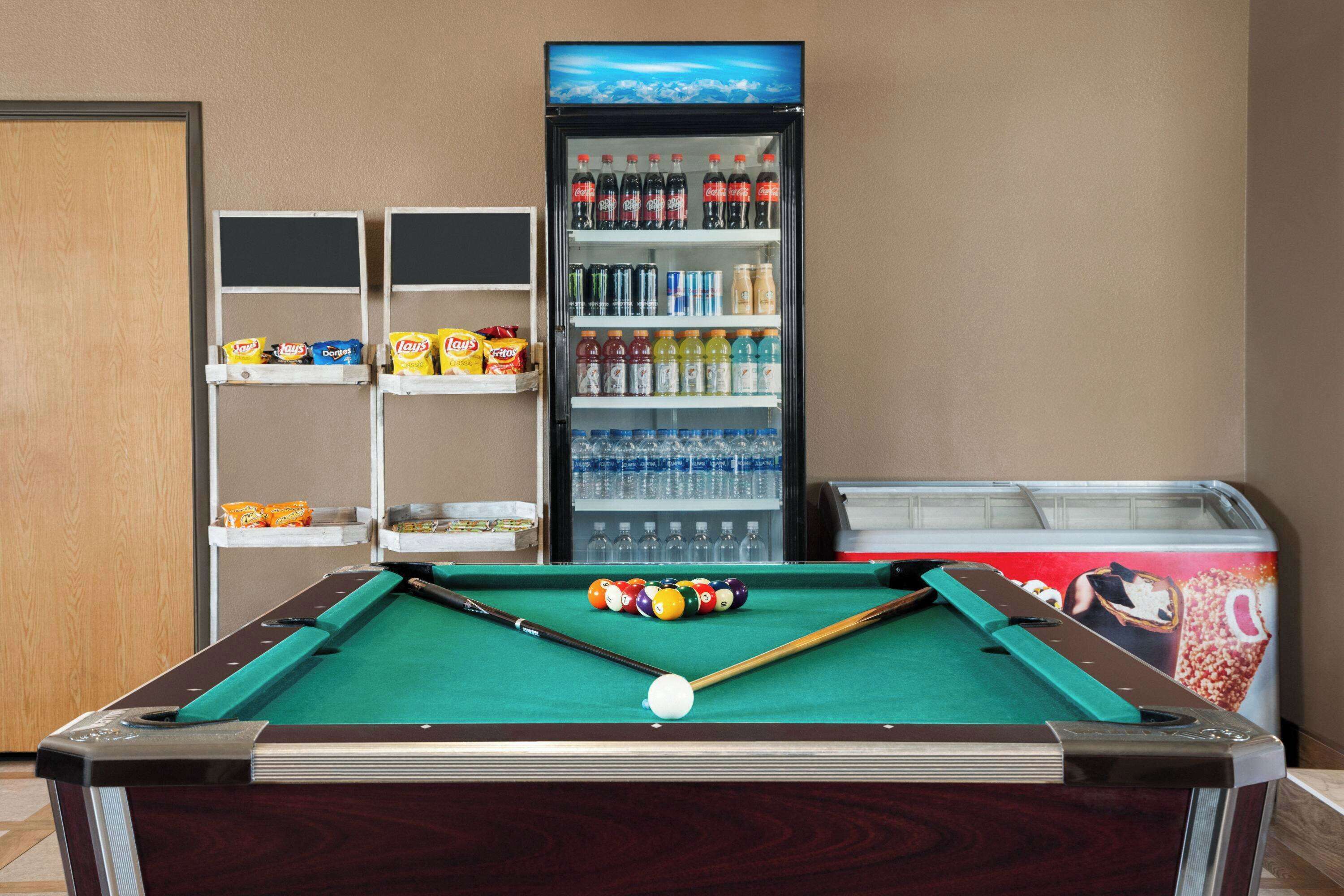 A pool table with billiard balls and cues, a vending machine with beverages, and snack shelves.