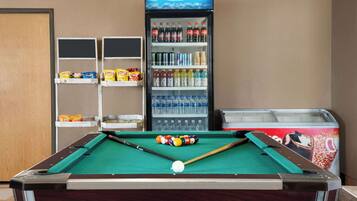 A pool table with billiard balls and cues, a vending machine with beverages, and snack shelves.