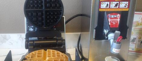 Una máquina para hacer gofres con un gofre dentro, un dispensador de carga directa y una botella de jarabe sobre una encimera.