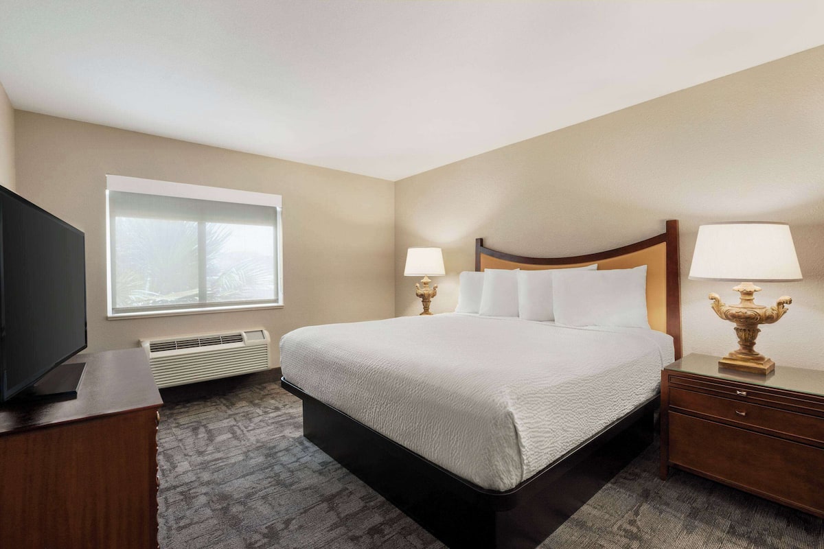 A hotel room with a large bed, two bedside lamps, a TV, and a window with blinds.