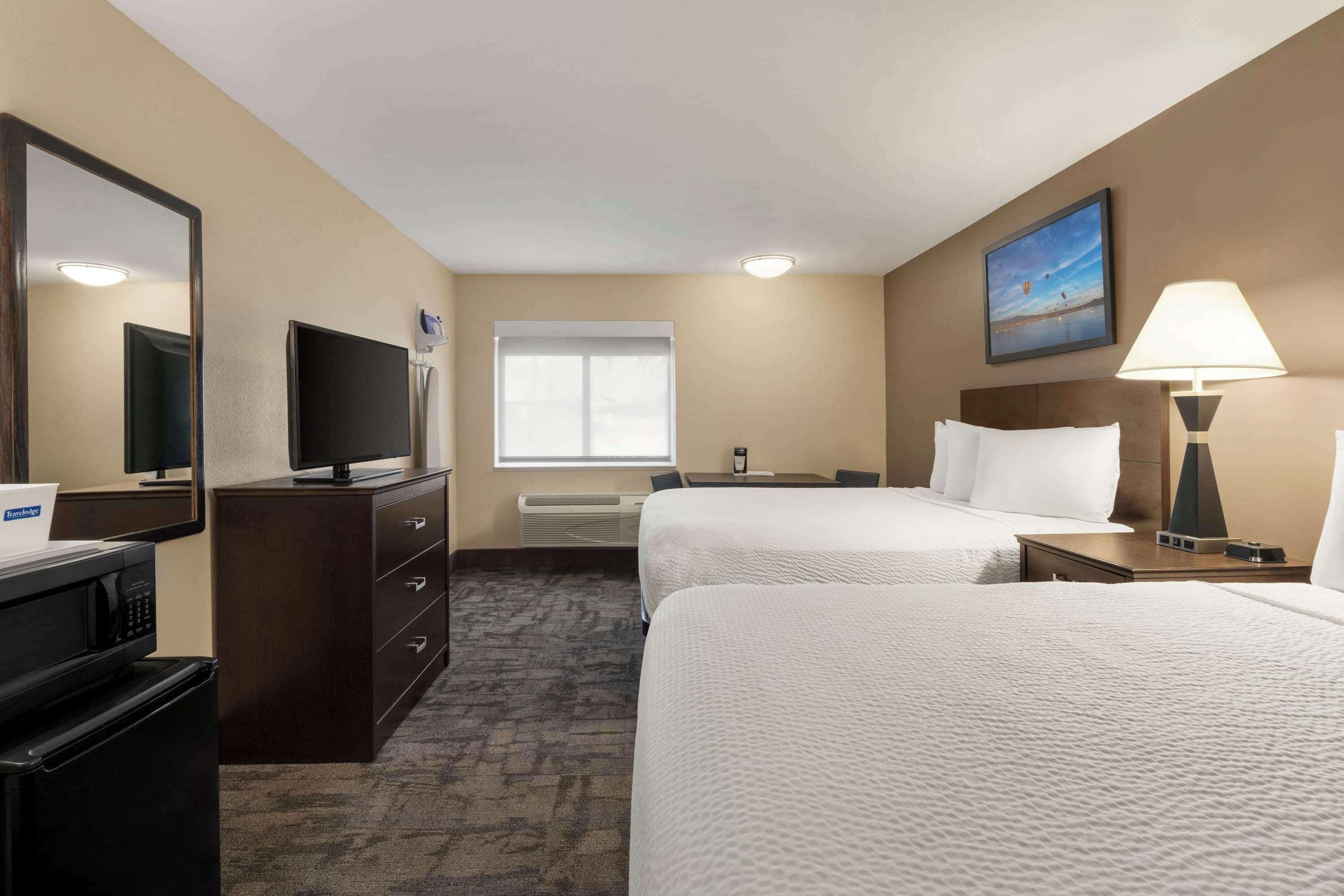 Habitación de hotel con dos camas, un escritorio, televisor, espejo, ventana y lámpara.