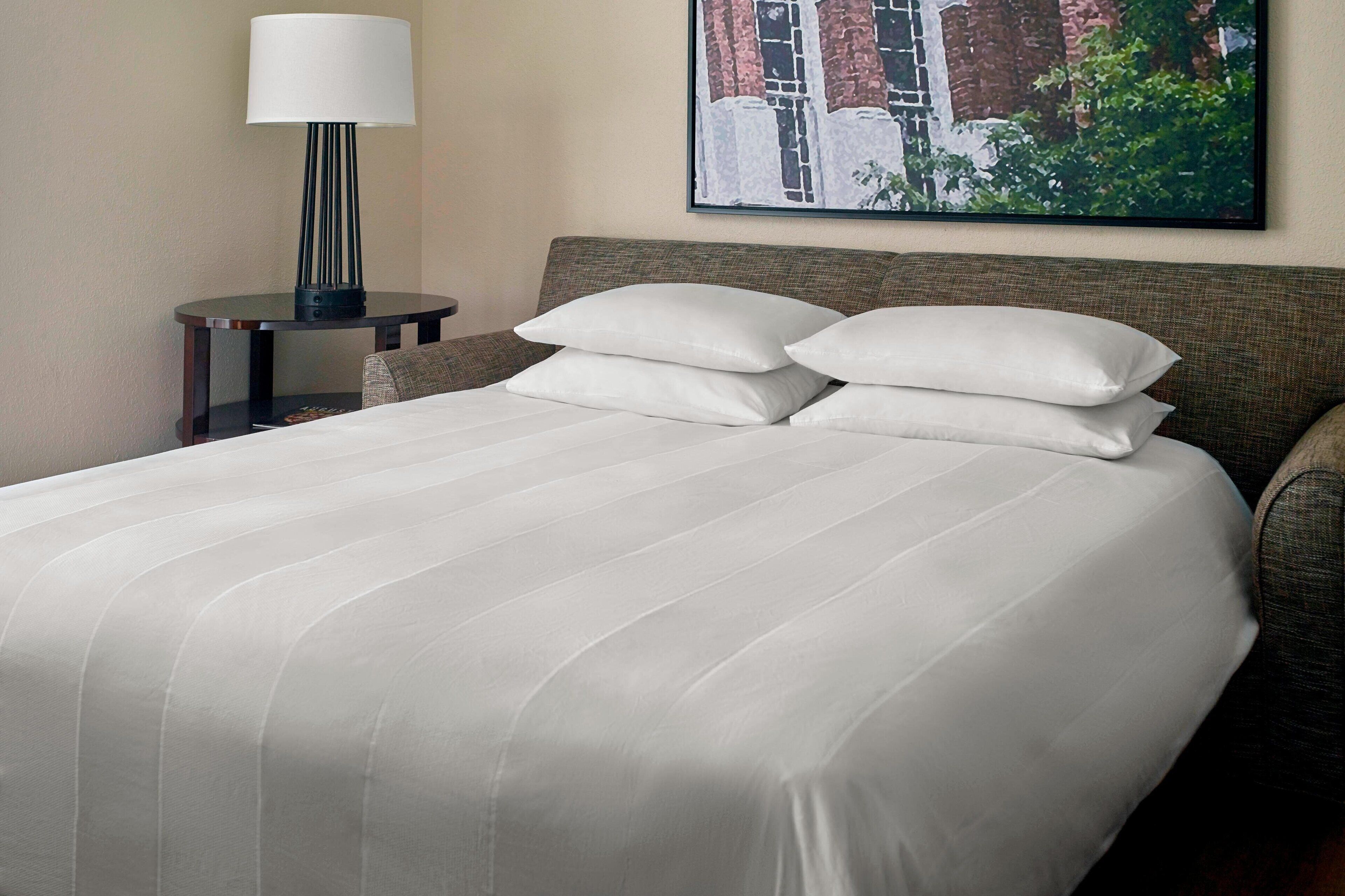 A neatly made bed with white linens, a bedside table with a lamp, and a framed picture on the wall.