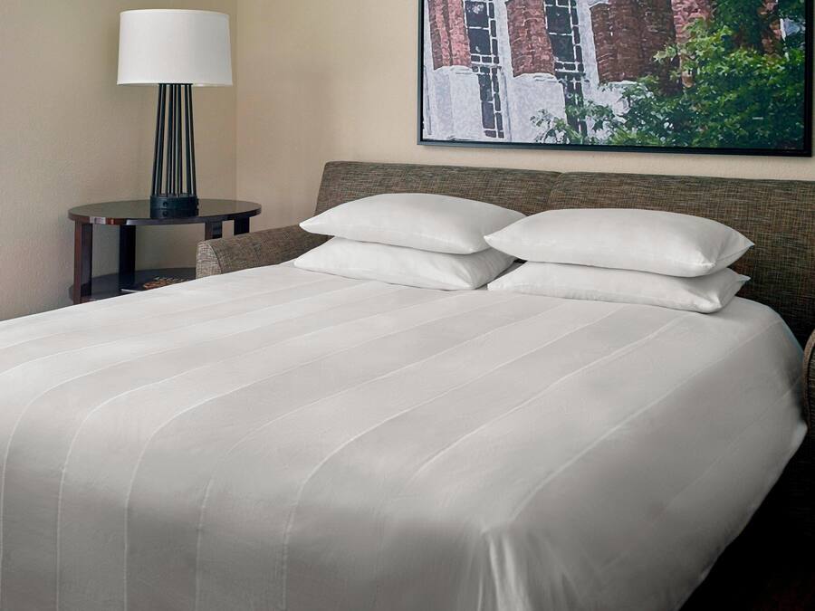 Una cama bien tendida con sábanas blancas, una mesita de noche con lámpara y un cuadro en la pared.