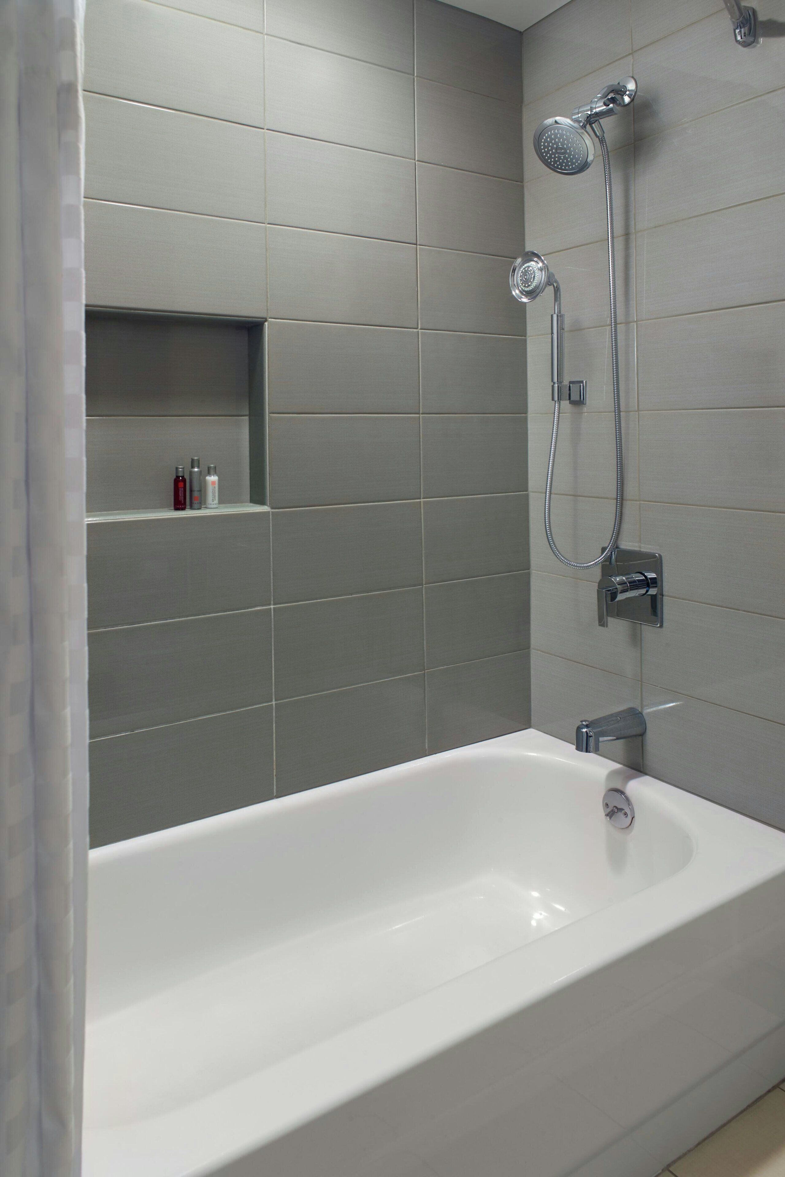 Un baño moderno con una bañera grande, ducha con cabezal de ducha manual y grifo empotrado en la pared. Hay dos estantes pequeños con artículos de tocador.