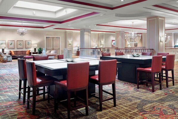 A hotel dining area with a large table, red upholstered chairs, chandeliers, and a patterned carpet.