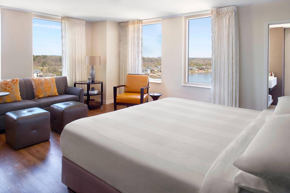 A hotel room with a large bed, a sofa, two ottomans, a chair, and a small table with a lamp.