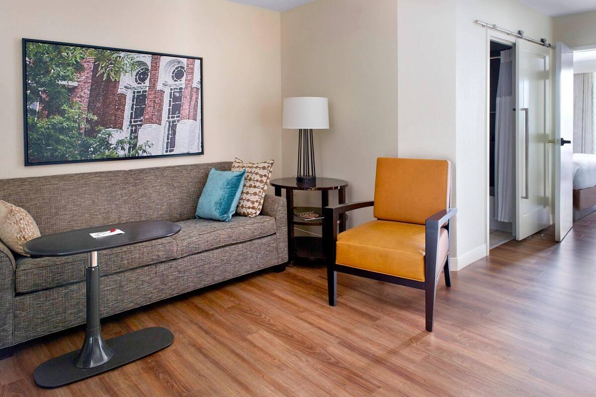 A modern hotel room with a sofa, a small table, a chair, a lamp, and a painting on the wall.