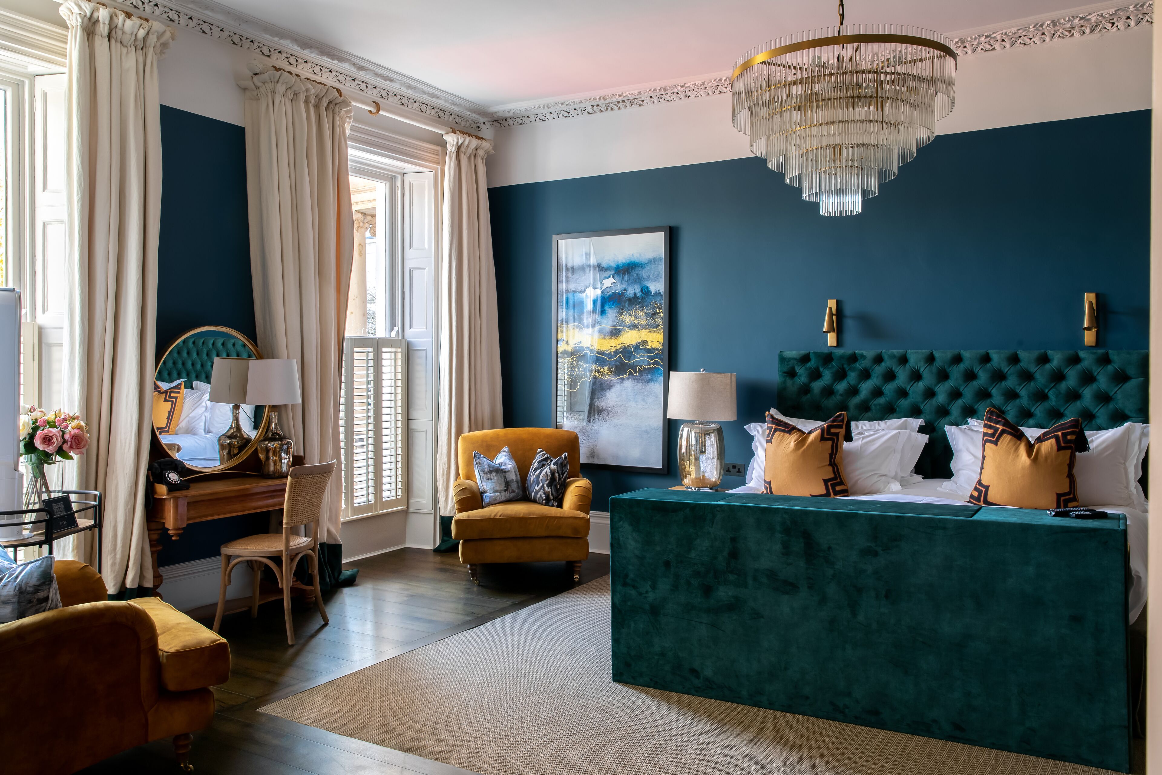 A room with a teal upholstered headboard, a yellow armchair, a wooden desk with a lamp, and a large window with curtains.