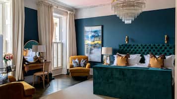 A room with a teal upholstered headboard, a yellow armchair, a wooden desk with a lamp, and a large window with curtains.