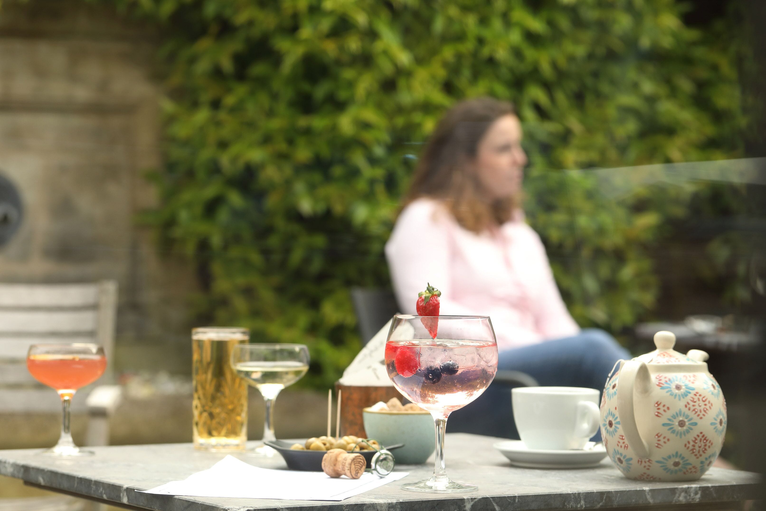 A table with a variety of drinks, a teapot, and a cup, set against a backdrop of greenery and a stone wall.