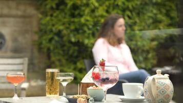 A table with a variety of drinks, a teapot, and a cup, set against a backdrop of greenery and a stone wall.