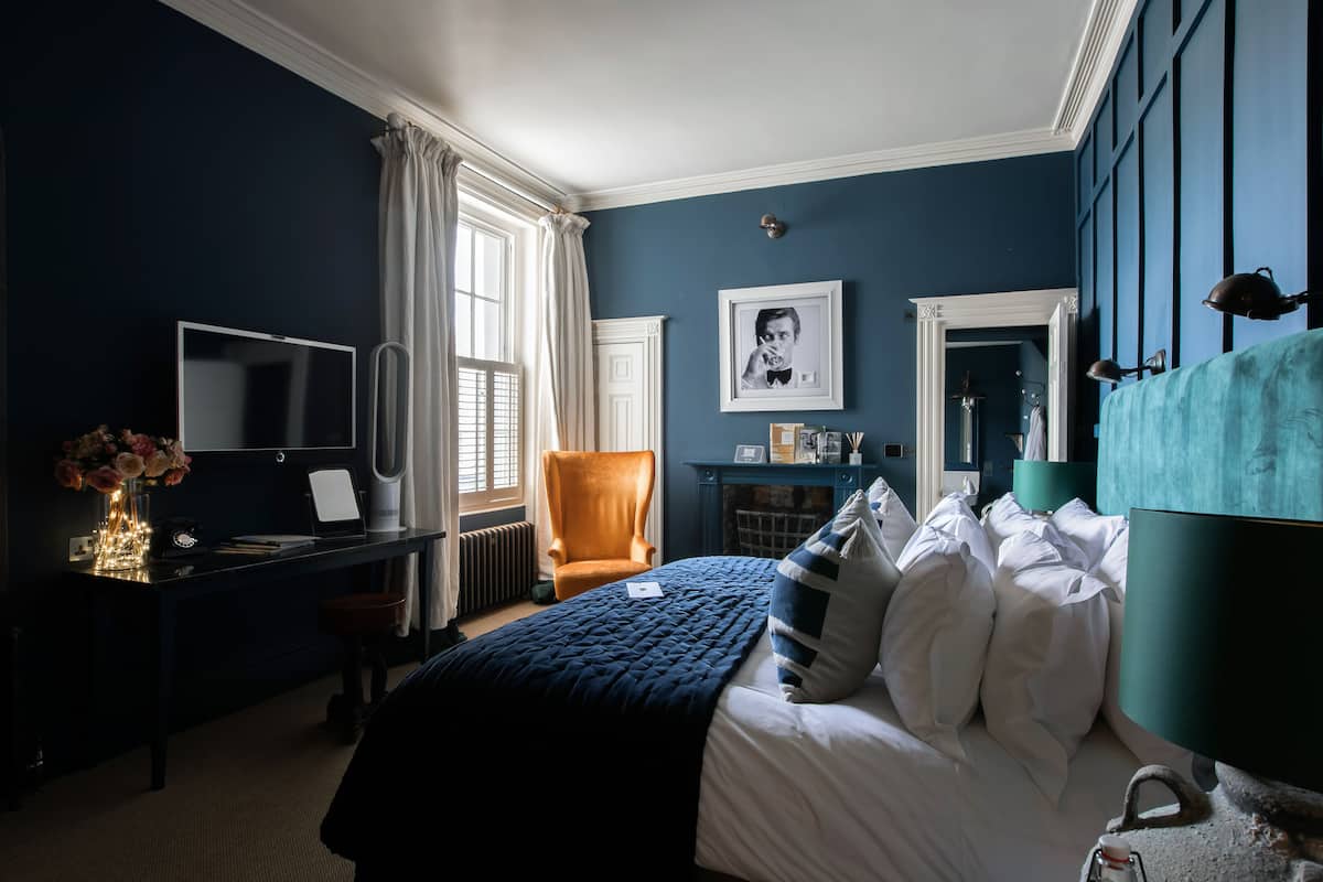 Habitación de hotel con pared azul oscuro, cama grande con ropa de cama blanca y azul, escritorio con computadora y un florero, una silla y una chimenea.