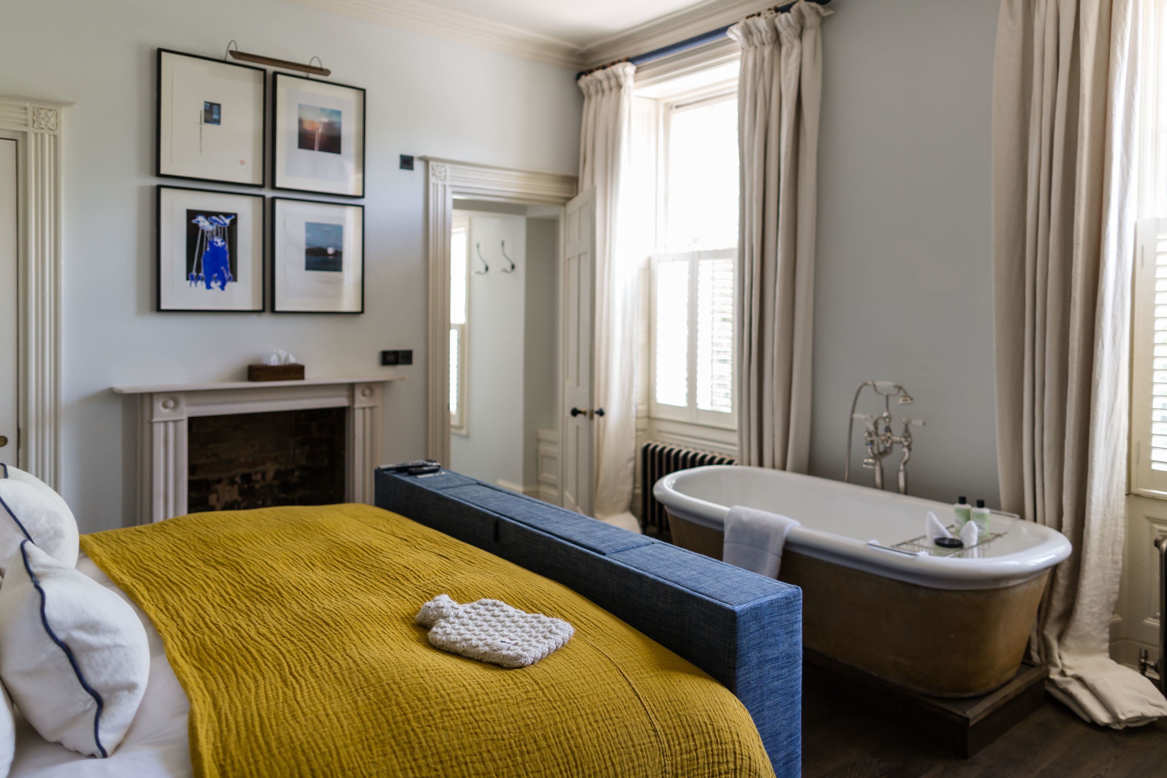 A bedroom with a bed, a blue bench, a fireplace, a bathtub, and framed pictures on the wall.