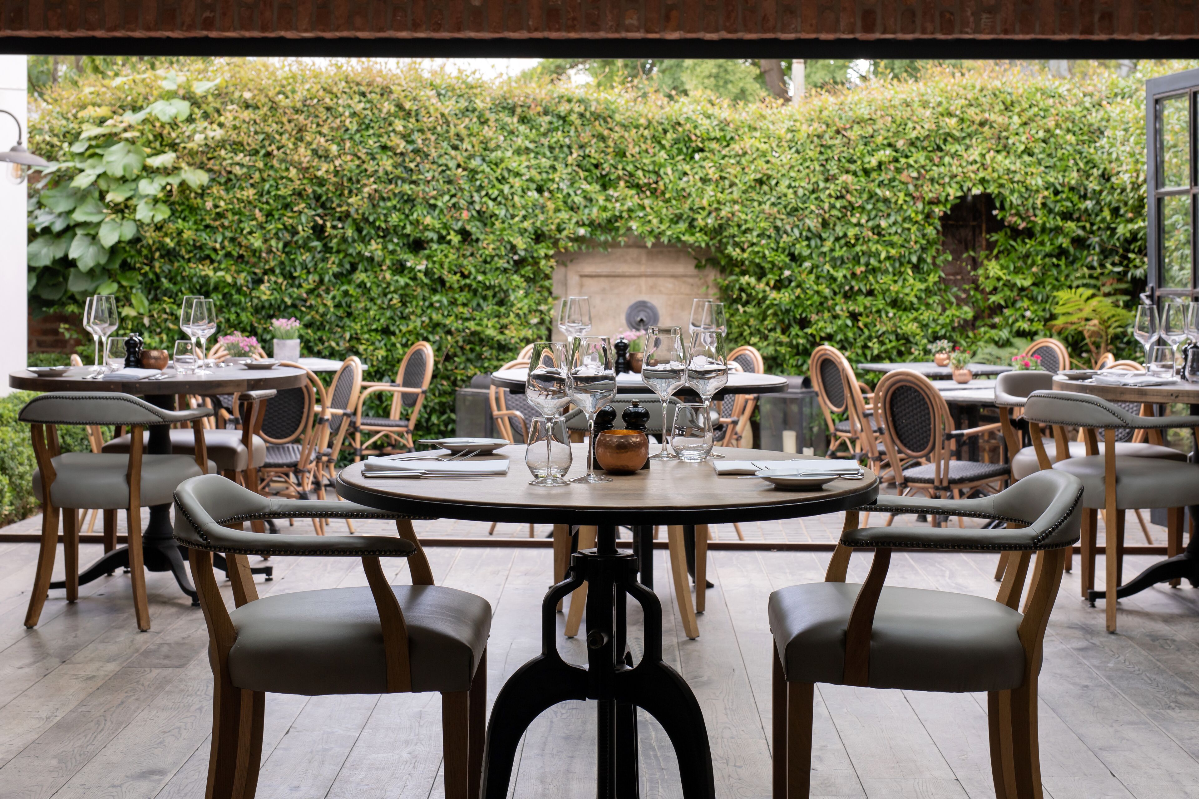 Un restaurant avec des tables rondes et des chaises rembourrées, entouré d’une haie verdoyante et luxuriante.