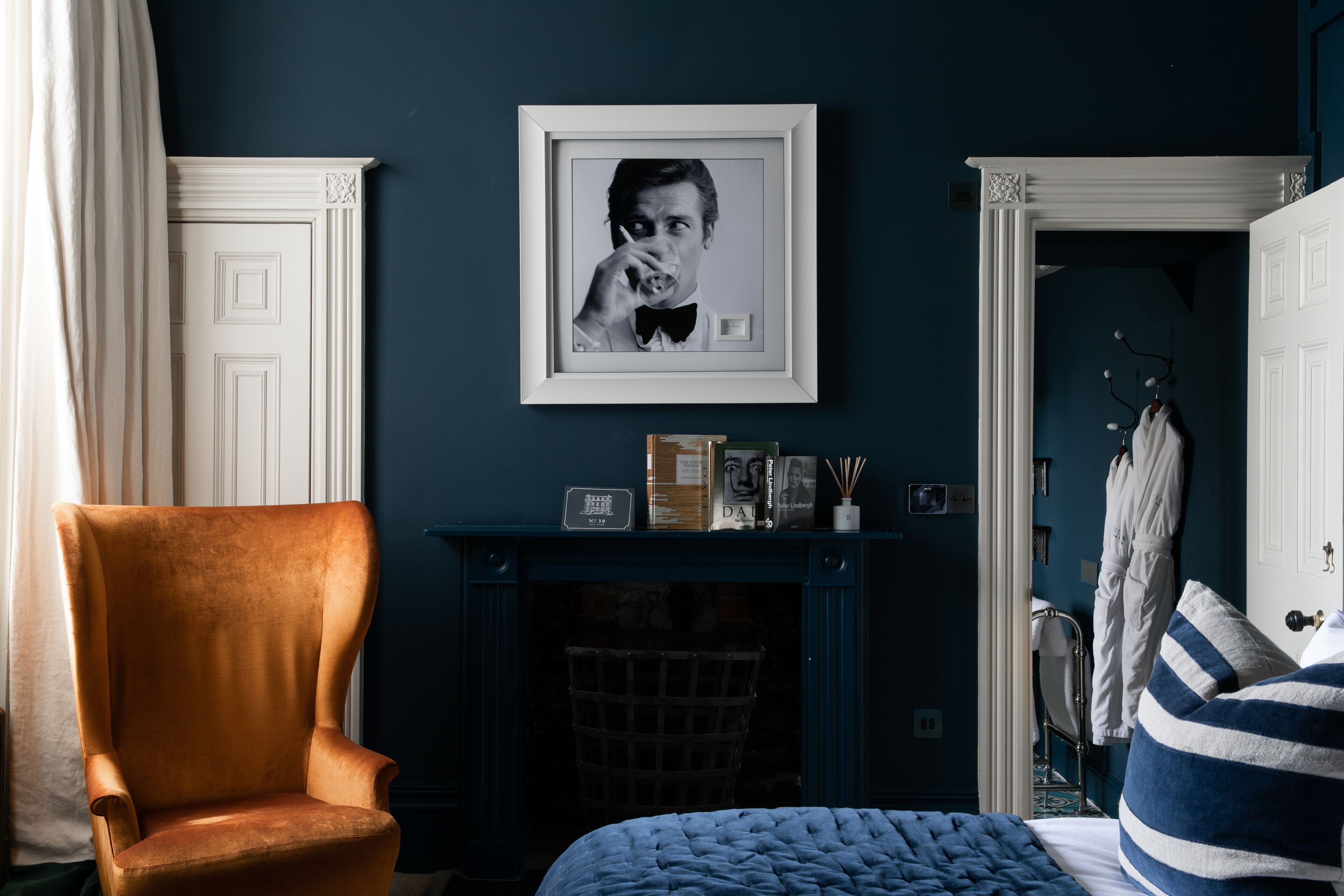 A room with a large armchair, a bed with a blue quilt, a framed picture on the wall, and a fireplace.