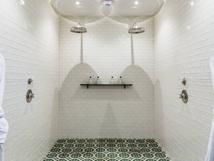 A shower with white tiled walls, a green and white patterned floor, and two overhead showerheads.